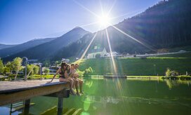 Eine Familie mit zwei Kindern sitzt gemeinsam auf einem Steg im Wasserpark See, während die Sonne auf die umliegenden Berge scheint.