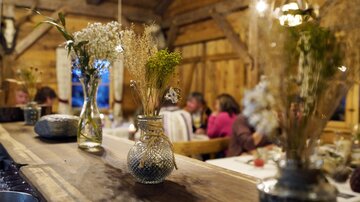 Eine gemütliche Holzstube mit einem langen Tisch und verschnörkelten Vasen, die mit Trockenblumen gefüllt sind. Im Hintergrund sind Gäste zu sehen, die sich unterhalten.