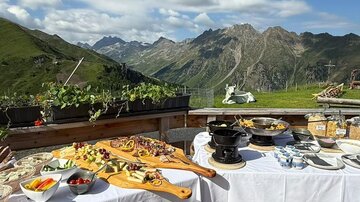 Ein gedeckter Tisch mit verschiedenen Speisen auf einer Terrasse in den Bergen. Im Hintergrund sind grüne Hügel und ein blauer Himmel zu sehen.
