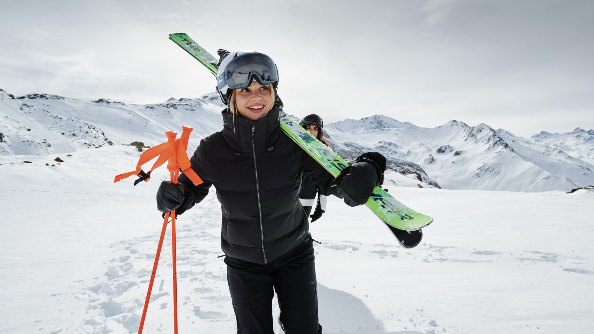 A skier glides down the slope with momentum. In the background, snow-covered mountains and a clear sky can be seen.