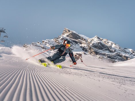 Ein Skifahrer fährt durch frischen Schnee in den Bergen. Im Hintergrund sind beeindruckende Felsen und ein blauer Himmel zu sehen.