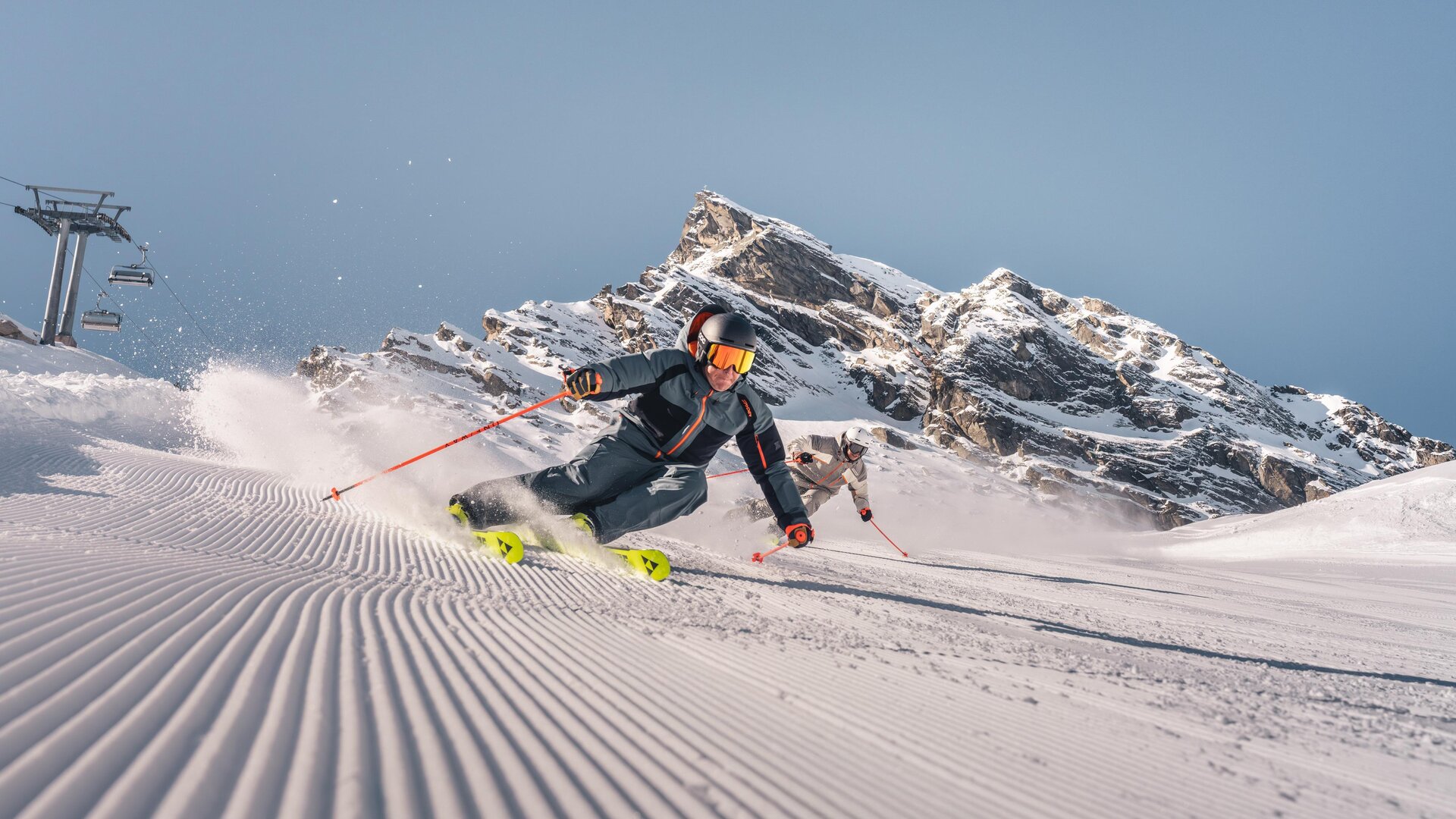 Ein Skifahrer fährt durch frischen Schnee in den Bergen. Im Hintergrund sind beeindruckende Felsen und ein blauer Himmel zu sehen.