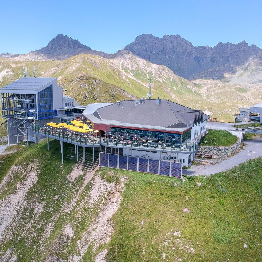 Ein modernes Bergrestaurant mit einer großen Terrasse, umgeben von grünen Wiesen und Bergen. Der klare Himmel bietet eine traumhafte Aussicht auf die alpine Landschaft.
