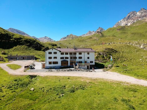 A mountain hut surrounded by green meadows and mountains. The sky is clear and blue.