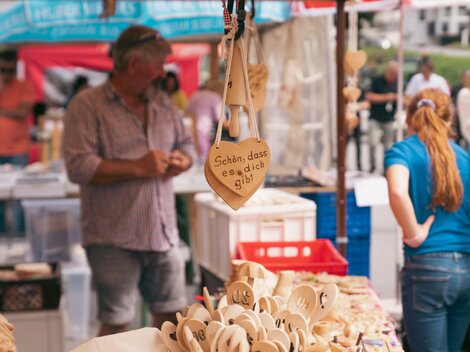 Ein Marktstand mit handgefertigten Holzartikeln. Im Hintergrund sind Menschen zu sehen, die das Angebot betrachten.