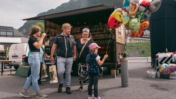 Eine Familie steht vor einem Verkaufsstand und betrachtet bunte Luftballons. Im Hintergrund sind grüne Berge zu sehen.