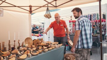 Ein Marktstand mit verschiedenen Holzobjekten ist zu sehen. Zwei Männer interagieren, während sie die Waren betrachten.