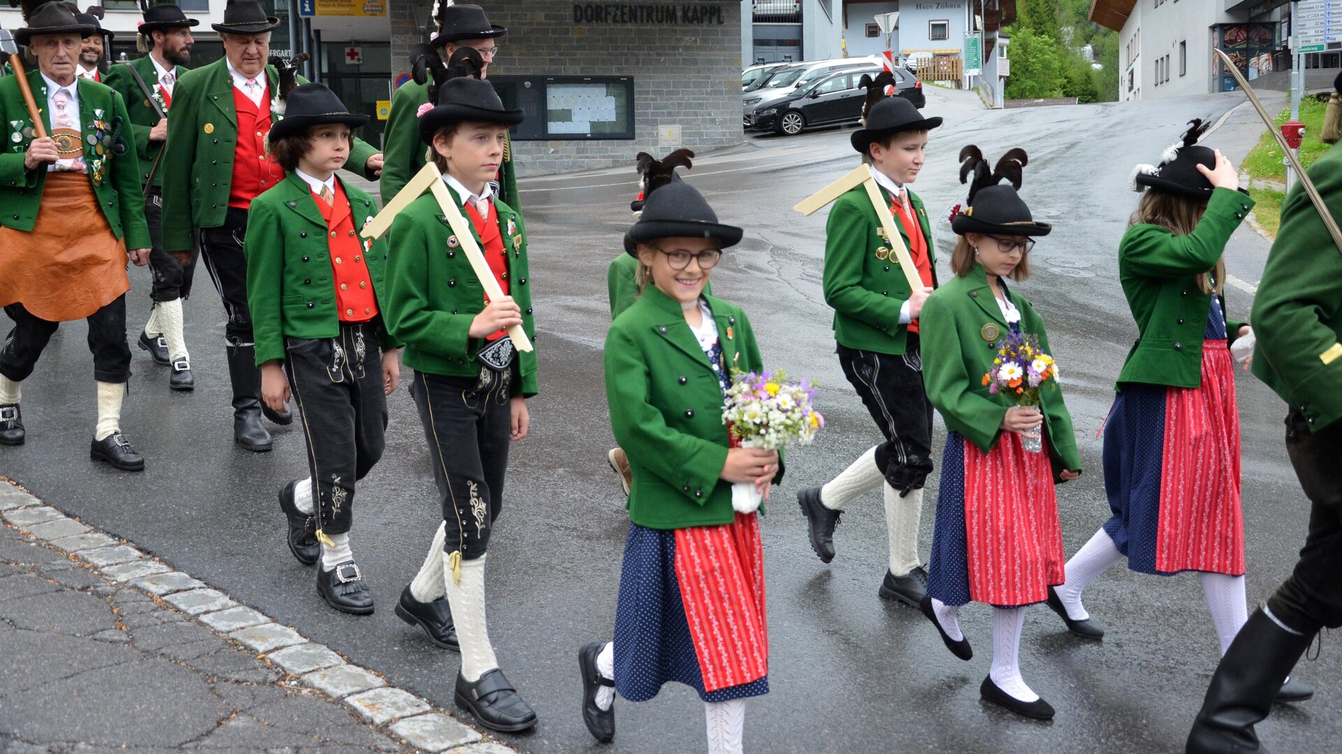 Eine Gruppe von traditionellen Trachtenträgern marschiert durch eine Straße. Sie tragen grüne Jacken und schwarze Hüte, und einige halten Blumen in den Händen.