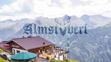 A mountain restaurant with a large terrace and a view of the surrounding mountains. The landscape is green and the sky is cloudy.