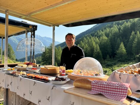 Ein freundlicher Verkäufer steht an einem Holzstand mit einer Vielzahl von Speisen. Im Hintergrund sind Berge und Wälder zu sehen.