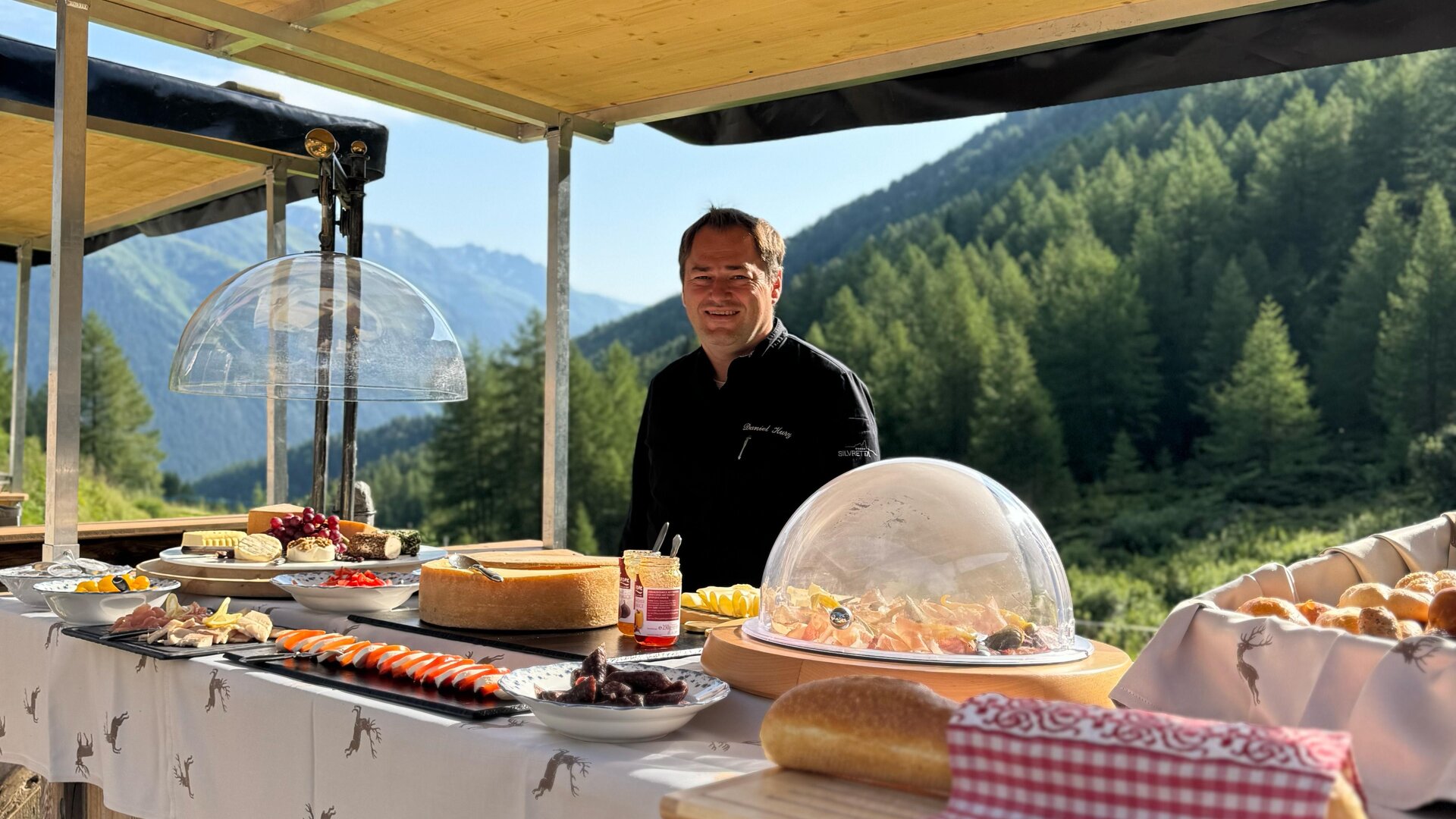 Ein freundlicher Verkäufer steht an einem Holzstand mit einer Vielzahl von Speisen. Im Hintergrund sind Berge und Wälder zu sehen.
