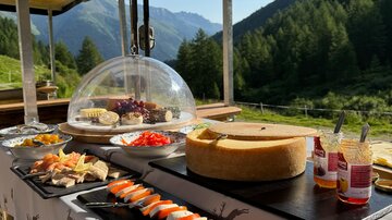 Ein festlich angerichteter Tisch im Freien mit einer Auswahl an Käse, Wurst und frischem Gemüse. Im Hintergrund erstrecken sich grüne Berge unter klarem Himmel.