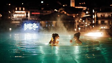Zwei Frauen genießen das Wasser in einem beleuchteten Pool bei Nacht. Im Hintergrund sind Lichter der Stadt sichtbar.