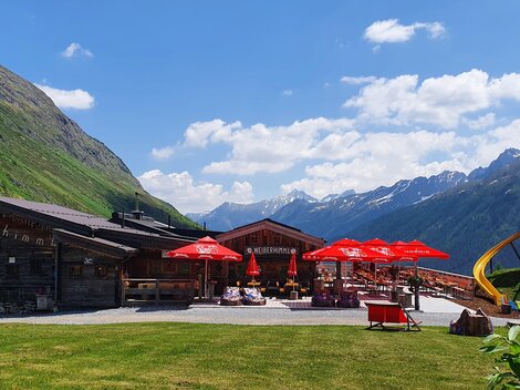 A cozy alpine village with red sun umbrellas and a playground. In the background, majestic mountains and blue sky can be seen.