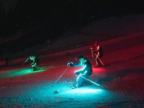 Drei Skifahrer mit leuchtenden, bunten Lichtern fahren im Dunkeln einen Hang hinunter. Die Umgebung ist schneebedeckt und die Stimmung ist festlich.