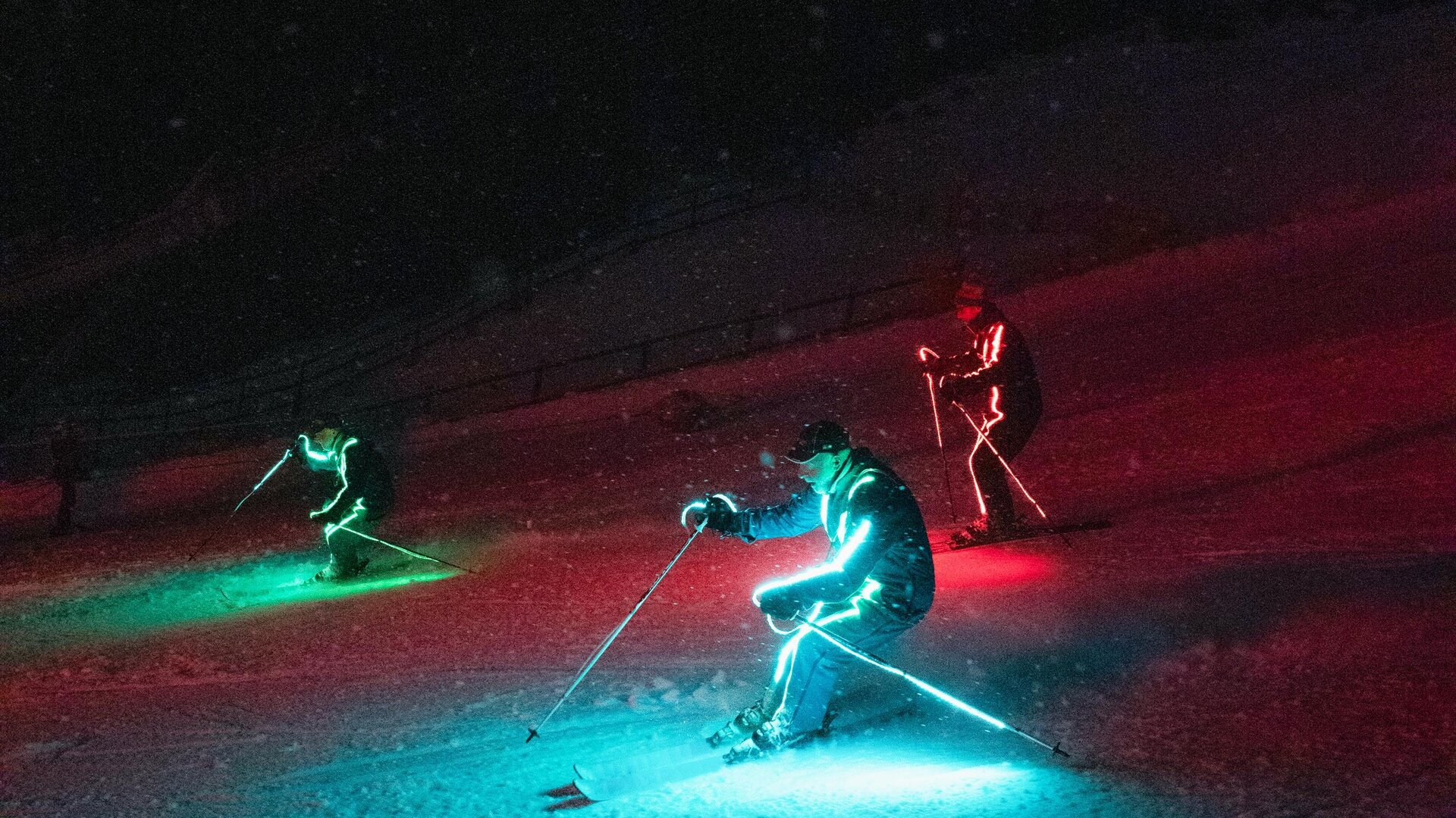 Drei Skifahrer mit leuchtenden, bunten Lichtern fahren im Dunkeln einen Hang hinunter. Die Umgebung ist schneebedeckt und die Stimmung ist festlich.
