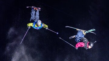 Zwei Skifahrer führen akrobatische Sprünge in der Luft aus. Der Hintergrund ist dunkel, und es ist sichtbar, wie Schnee aufgewirbelt wird.