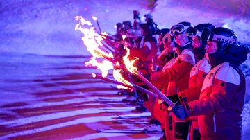 Eine Gruppe von Skifahrern in roten Anzügen steht an einer beleuchteten Piste mit Fackeln. Die Flammen erhellen die Winterlandschaft und schaffen eine festliche Atmosphäre.