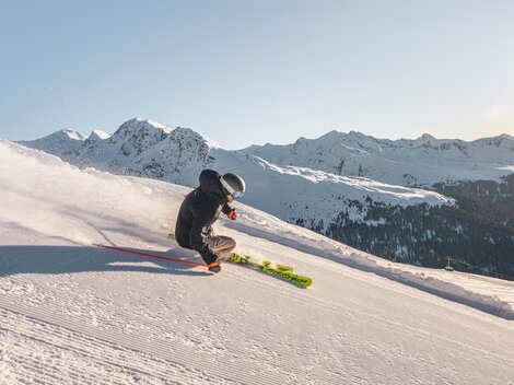 Ein Skifahrer fährt die schneebedeckte Piste hinunter. Im Hintergrund sind majestätische Berge zu sehen.