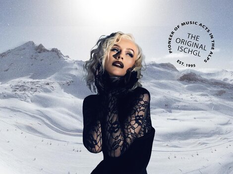 A glamorous model poses in front of a snow-covered mountain landscape. Majestic mountains and a clear sky are visible in the background.