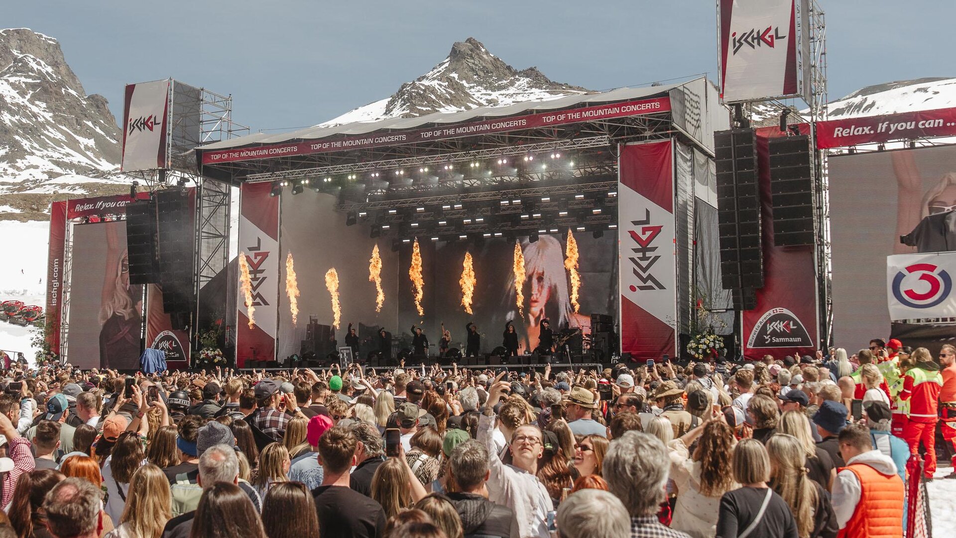 A large music festival in the mountains with an impressive stage. Many people enjoy the performance under clear skies.