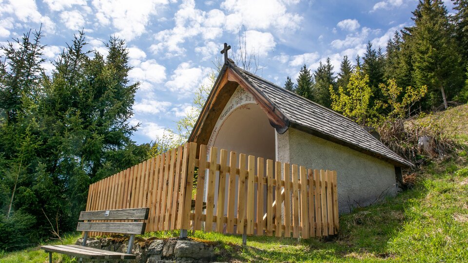 Eine kleine Kapelle mit einem Holzzaun steht in einer bewaldeten Umgebung. Der Himmel ist teilweise bewölkt und die Landschaft wirkt ruhig und friedlich. | © Bernhard Gruber – Photography