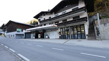 Ein Gebäude mit einer Tankstelle und Verkaufsräumen an einer ruhigen Straße. Im Hintergrund sind weitere traditionelle alpine Gebäude sichtbar.