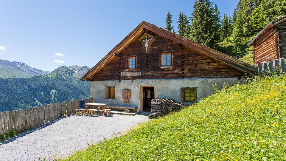 Ein traditionelles Holzhaus in den Bergen, umgeben von grünen Wiesen und Bäumen. Die klare, sonnige Landschaft bietet einen wunderschönen Ausblick auf die umliegenden Berge.