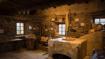 Eine gemütliche und rustikale Innenansicht eines alten Bauernhauses. Zu sehen sind Holz- und Steinstrukturen sowie landwirtschaftliche Werkzeuge.
