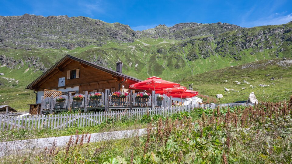 Eine malerische Almhütte umgeben von grünen Wiesen und Bergen. Rote Sonnenschirme stehen vor der Terrasse, die einladend wirkt.