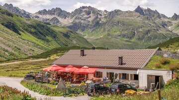 Eine gemütliche Hütte in den Bergen mit roten Sonnenschirmen. Die Umgebung ist grün und bergig, ideal für Naturfreunde.