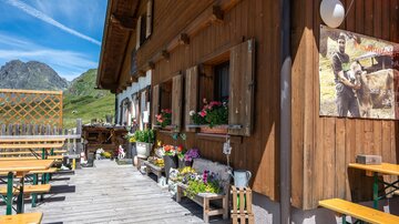 Eine charmante Berghütte mit Holzwänden und blühenden Blumen. Draußen stehen Tische und Bänke auf einer Holzterrasse.