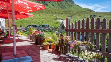 Ein gemütlicher Außenbereich mit roten Sonnenschirmen und blühenden Pflanzen. Im Hintergrund sind grüne Berge und einige Autos zu sehen.