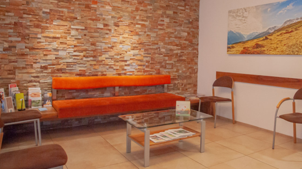 Ein modernes Wartezimmer mit einer orangen Bank und Braunstuhl. Die Wand ist mit künstlichen Steinen verkleidet, und ein Bild einer Landschaft hängt darüber.