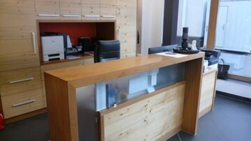 A modern reception area with a wooden desk and light-colored walls. In the background, there are office furniture and large windows that let in light.