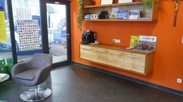 A modern reception area with an orange wall and a wooden shelving wall. The room features a gray armchair and a coffee machine.