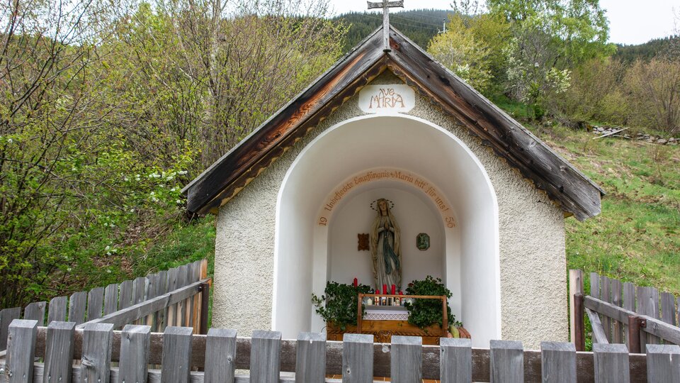 Eine kleine Kapelle mit einem heiligen Standbild steht in der Natur. Sie ist von einem Holzzaun umgeben und von Bäumen umgeben. | © Bernhard Gruber – Photography