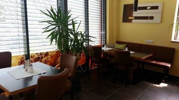 Ein modernes Restaurant mit einer gemütlichen Atmosphäre. Große Fenster lassen viel Licht herein, und Pflanzen sorgen für ein angenehmes Ambiente.