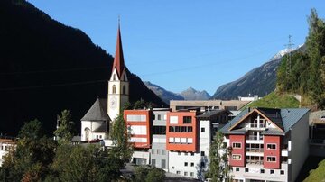 Ein malerisches Dorf mit einer Kirche und modernen Gebäuden. Im Hintergrund sind Berglandschaften zu sehen.