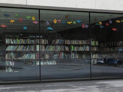 Ein modernes Fenster mit bunten Tiermotiven, das auf eine Bücherwand blickt. Die Bücher sind ordentlich in Regalen angeordnet.