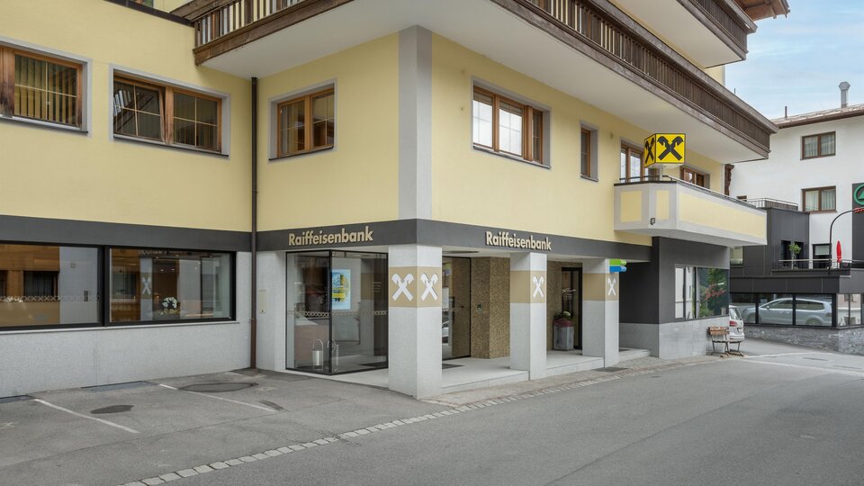 Ein modernes Bankgebäude mit gelber Fassade und großen Fenstern. Vor dem Gebäude befindet sich ein Parkplatz und eine Basketballanlage.
