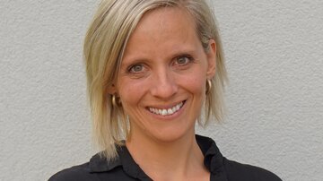 Eine lächelnde Frau mit blonden Haaren steht vor einer hellen Wand. Sie trägt ein schwarzes Oberteil und sieht freundlich aus.