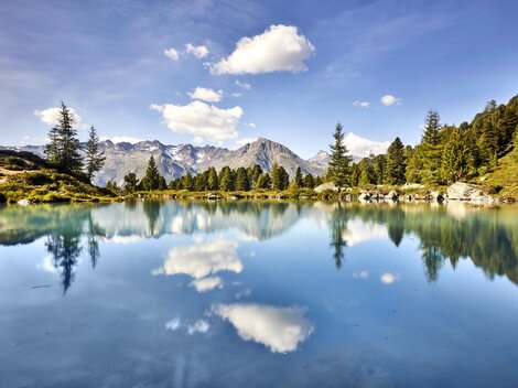 Ein ruhiger Bergsee mit kristallklarem Wasser und spiegelnden Wolken. Die umliegenden Bäume und Berge schaffen eine idyllische Naturszene.