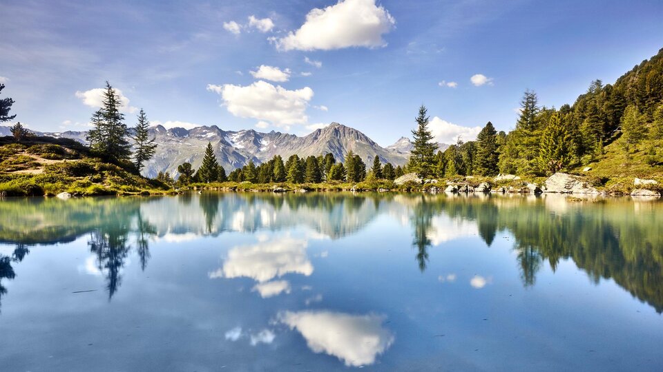 Ein ruhiger Bergsee mit kristallklarem Wasser und spiegelnden Wolken. Die umliegenden Bäume und Berge schaffen eine idyllische Naturszene.