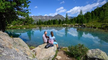 Ein ruhiger See umgeben von Bergen und Bäumen. Zwei Personen sitzen am Ufer und genießen die Aussicht.