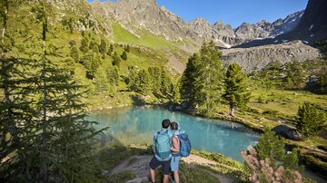 Ein Paar steht an einem klaren See in einer schönen Berglandschaft. Die umliegenden Berge sind grün und sonnig.