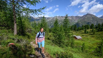 Eine Frau wandert auf einem Pfad durch eine grüne Landschaft. Im Hintergrund sind Berge und eine kleine Hütte zu sehen.