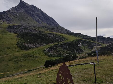 Eine grüne Berglandschaft mit einem hohen Gipfel im Hintergrund. Im Vordergrund steht ein Hinweis- oder Informationsschild.