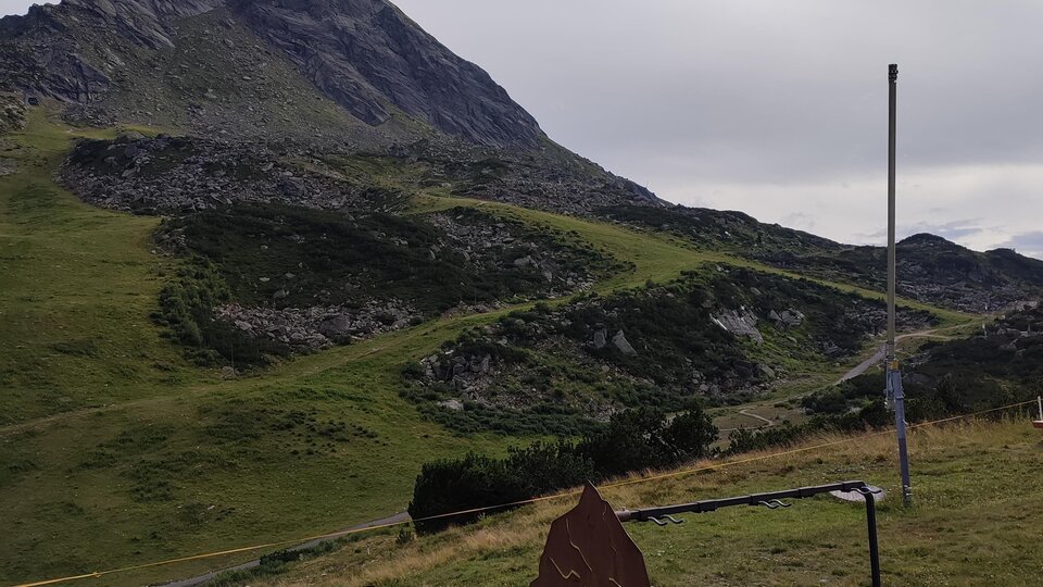 Eine grüne Berglandschaft mit einem hohen Gipfel im Hintergrund. Im Vordergrund steht ein Hinweis- oder Informationsschild.