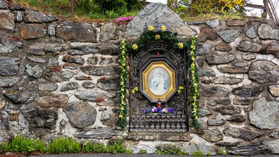 Eine Steinmauer mit einem dekorierten Bildnis in einer Nische. Das Bild ist mit Blumen geschmückt und zeigt eine religiöse Szene. | © Bernhard Gruber – Photography